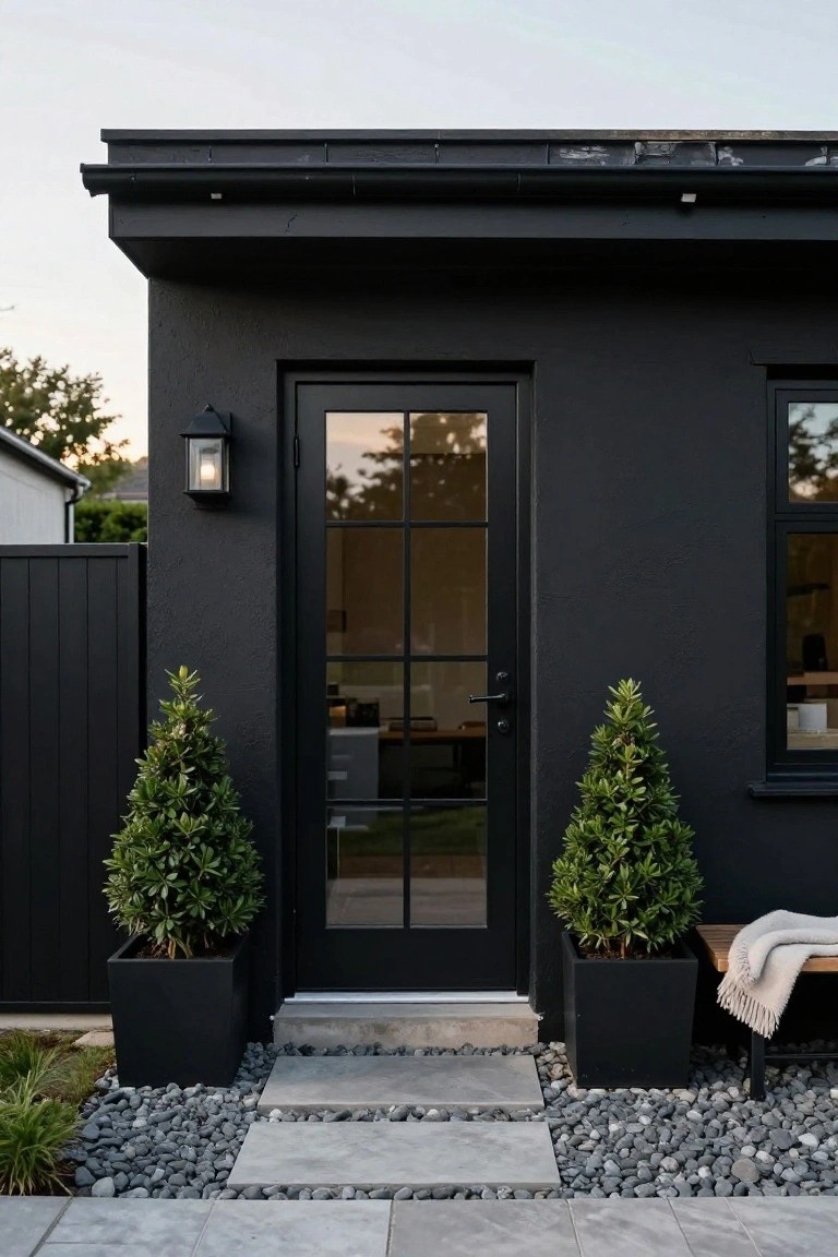 Matte black stucco bungalow exterior featuring a tall black steel-framed glass door with grid panes, flanked by two black pots of topiary trees, a wooden bench with throw, lantern light, and a gray pebble and paver pathway.