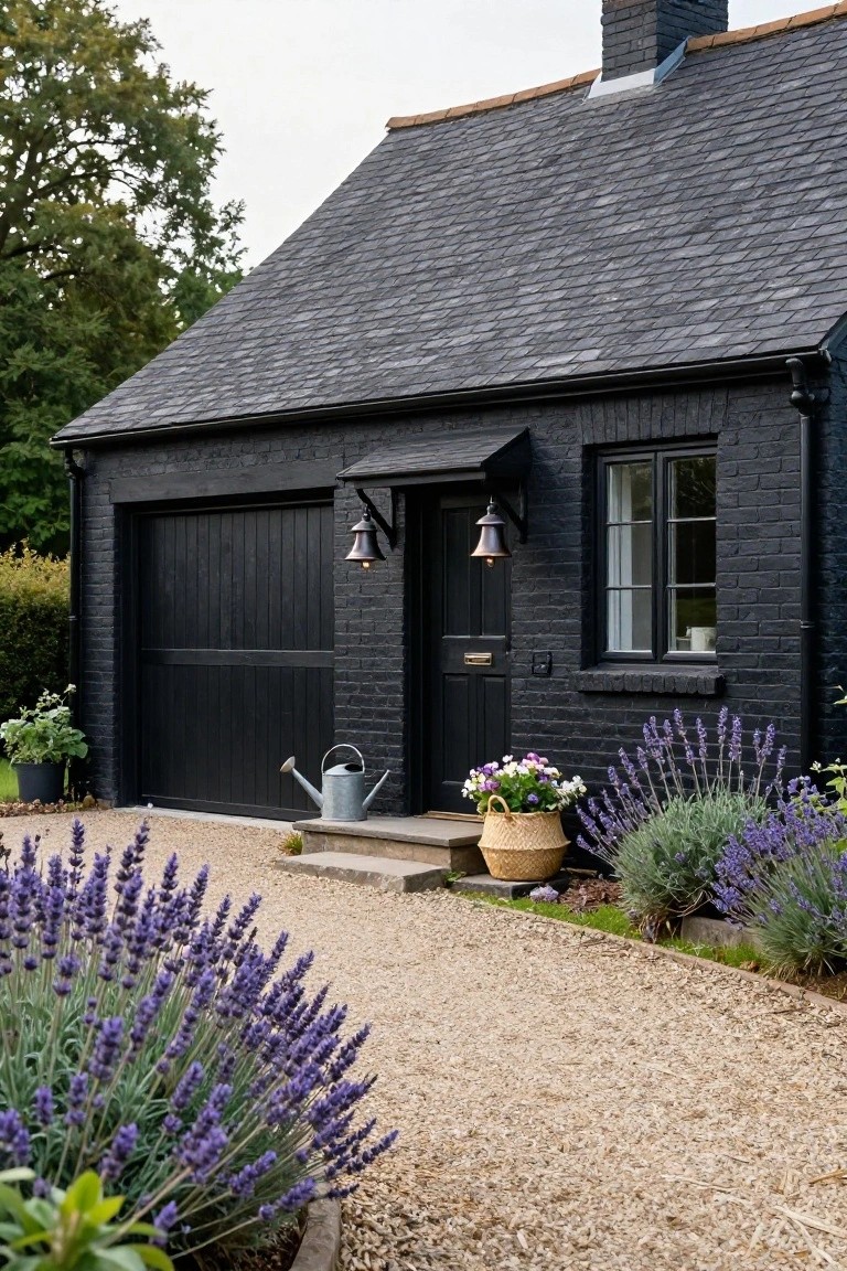 Black brick house exterior featuring a black garage door, black front door with lanterns, window, gravel path, lavender plants in beds, and a metal watering can near stone steps.