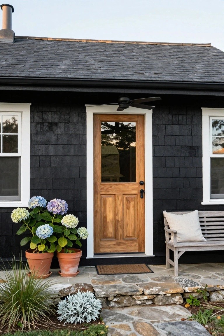 Black shingle house exterior with white window trim, glass-paneled wooden front door, potted blue hydrangeas, wooden bench with cushion, and irregular natural stone steps leading to the entryway.