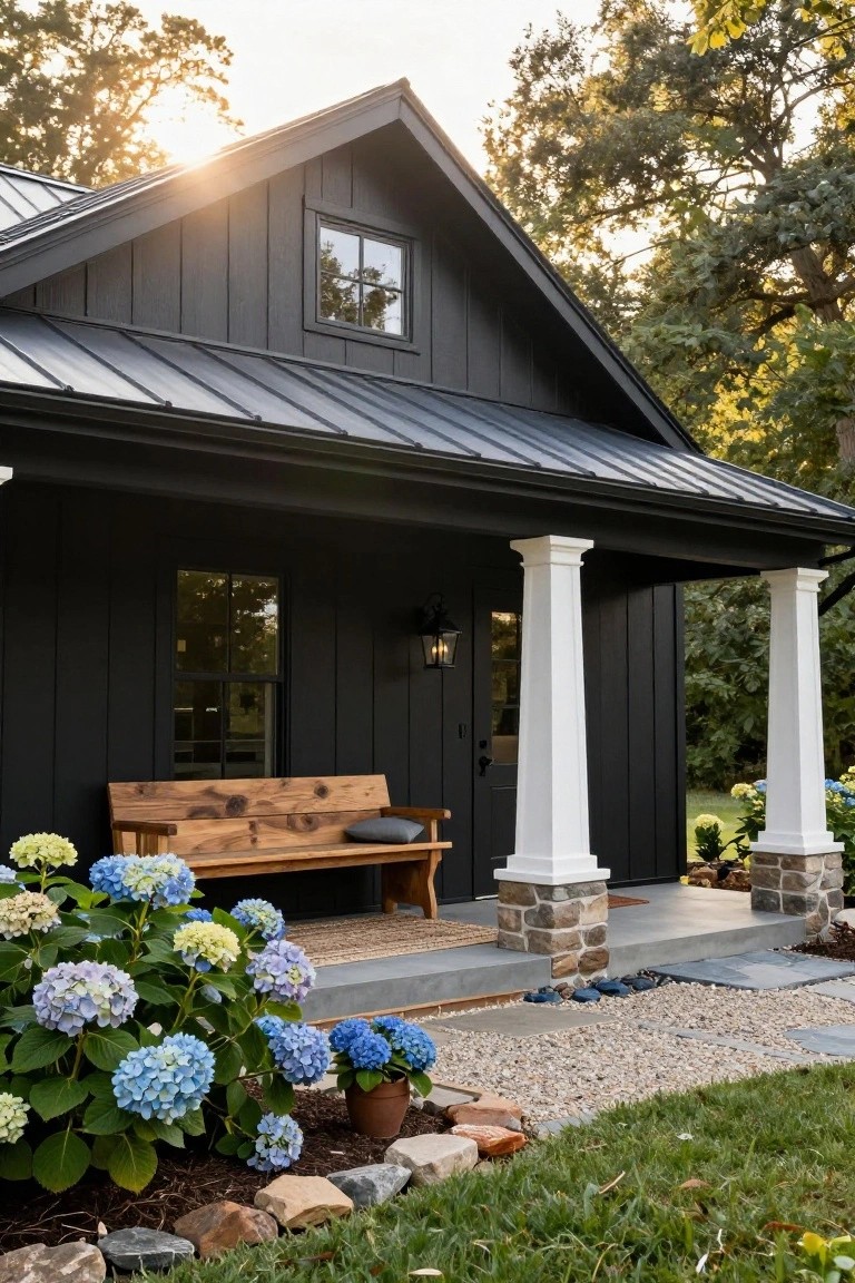 Black board-and-batten house exterior with metal roof, white porch columns on stone bases, wooden bench on porch, blue hydrangea bushes, and gravel path nearby.