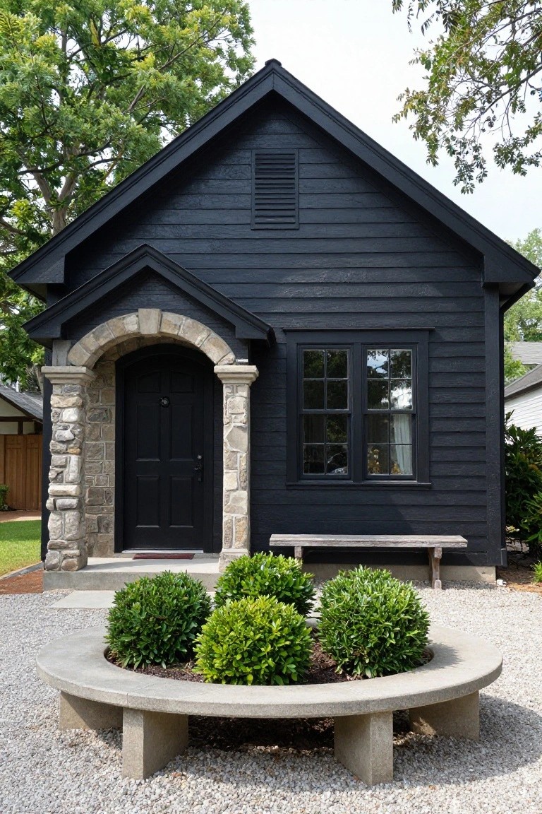 A small black shingled ranch-style house with a stone arched entryway featuring a dark wood door, a multipane window on the side, boxwood shrubs in a circular concrete planter on gravel, and surrounding trees and lawn.