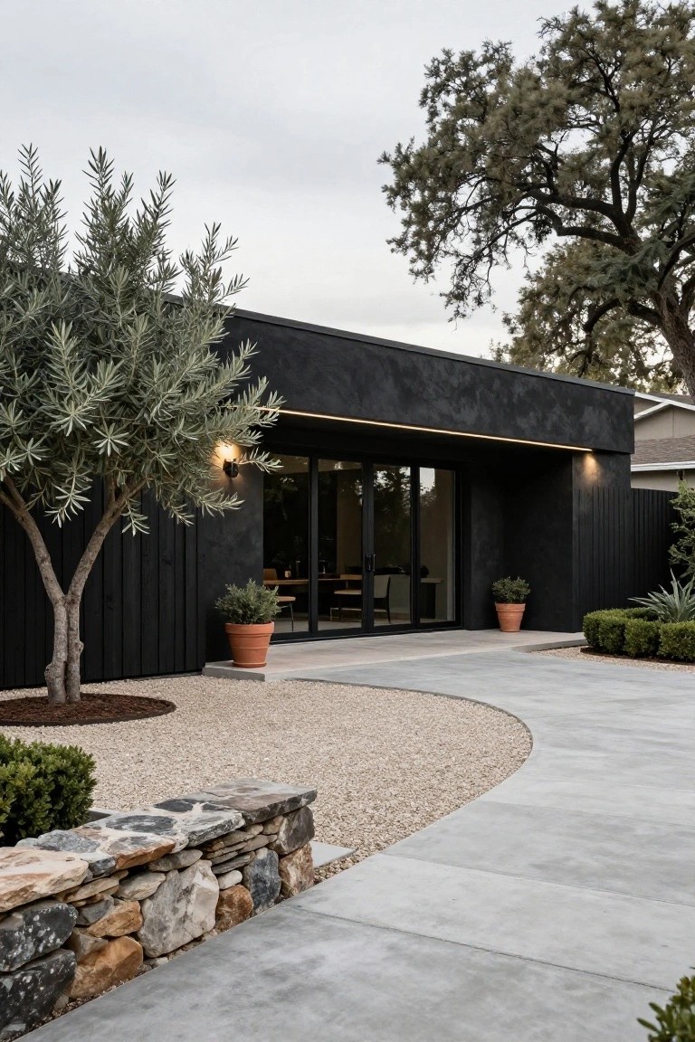 Black textured ranch house exterior with sliding glass entry doors, olive tree, potted plants in terracotta pots, gravel pathway, stone retaining wall, and surrounding landscaping.