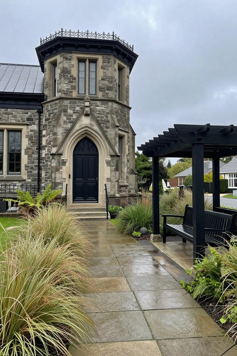 Gray stone Victorian house exterior featuring an octagonal tower with black top, tall pointed arch black double doors at the entrance, paver path lined with ornamental grasses leading to it, black pergola with bench nearby, under overcast sky.