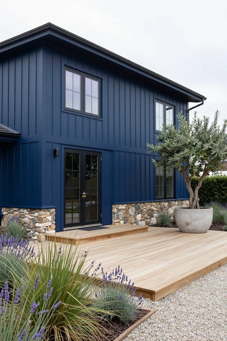 Dark blue vertical board-and-batten house exterior with stone foundation, wooden deck steps leading to black French doors, surrounded by gravel ground cover, lavender plants, ornamental grasses, and a large potted olive tree.