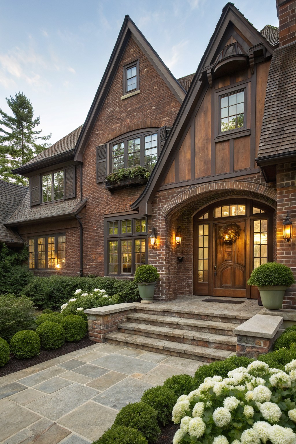 Two-story brick house exterior featuring dark brown brick walls, dark timber framing, arched wooden front door with lanterns, brick steps, and low shrubs with white flowers in the foreground.