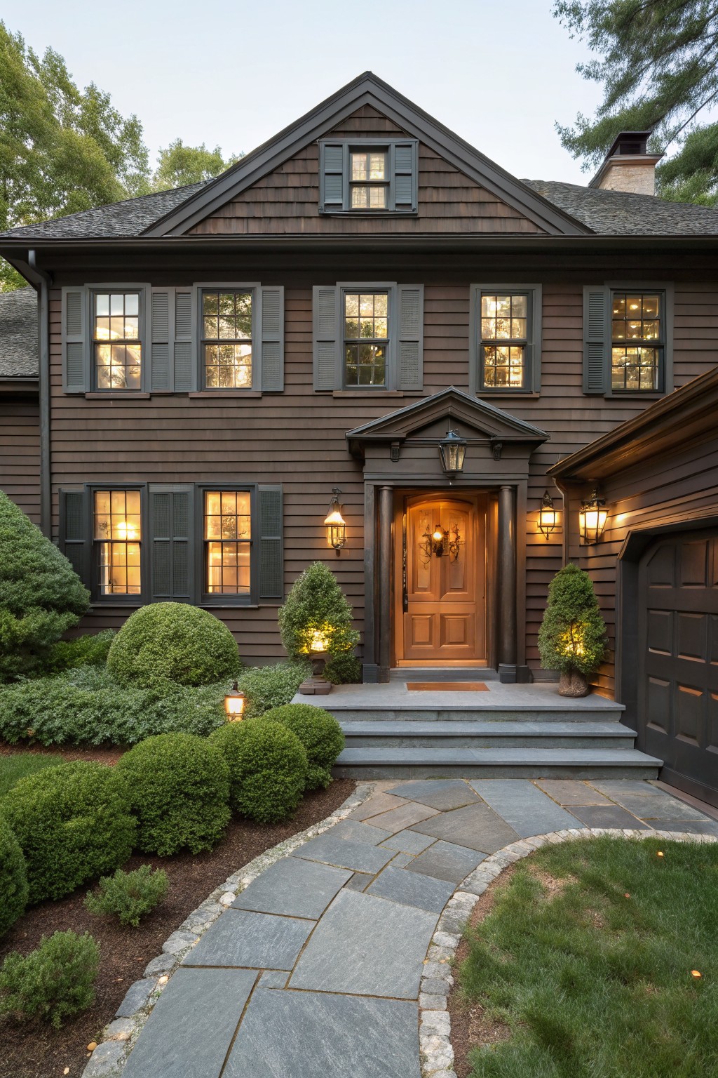 Front view of a two-story dark brown shingled house with gabled roof, dark shutters on lit windows, wooden arched door with lanterns, topiary shrubs, and curved stone pathway.