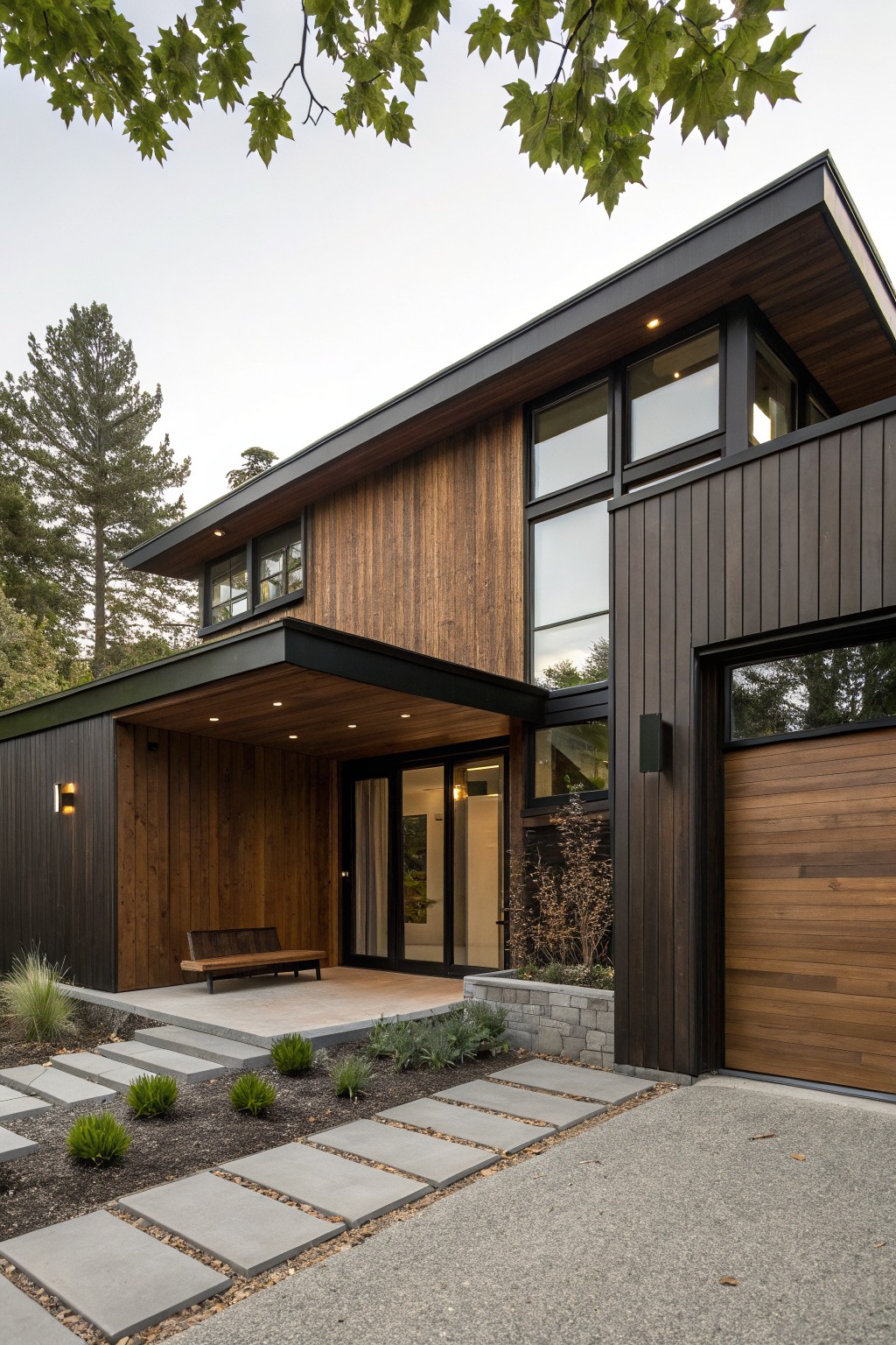 Modern two-story house exterior with dark brown vertical wood cladding, black metal accents, large windows, covered entry porch with bench, wooden garage door, stone steps, gravel driveway, and surrounding pine trees and grasses.