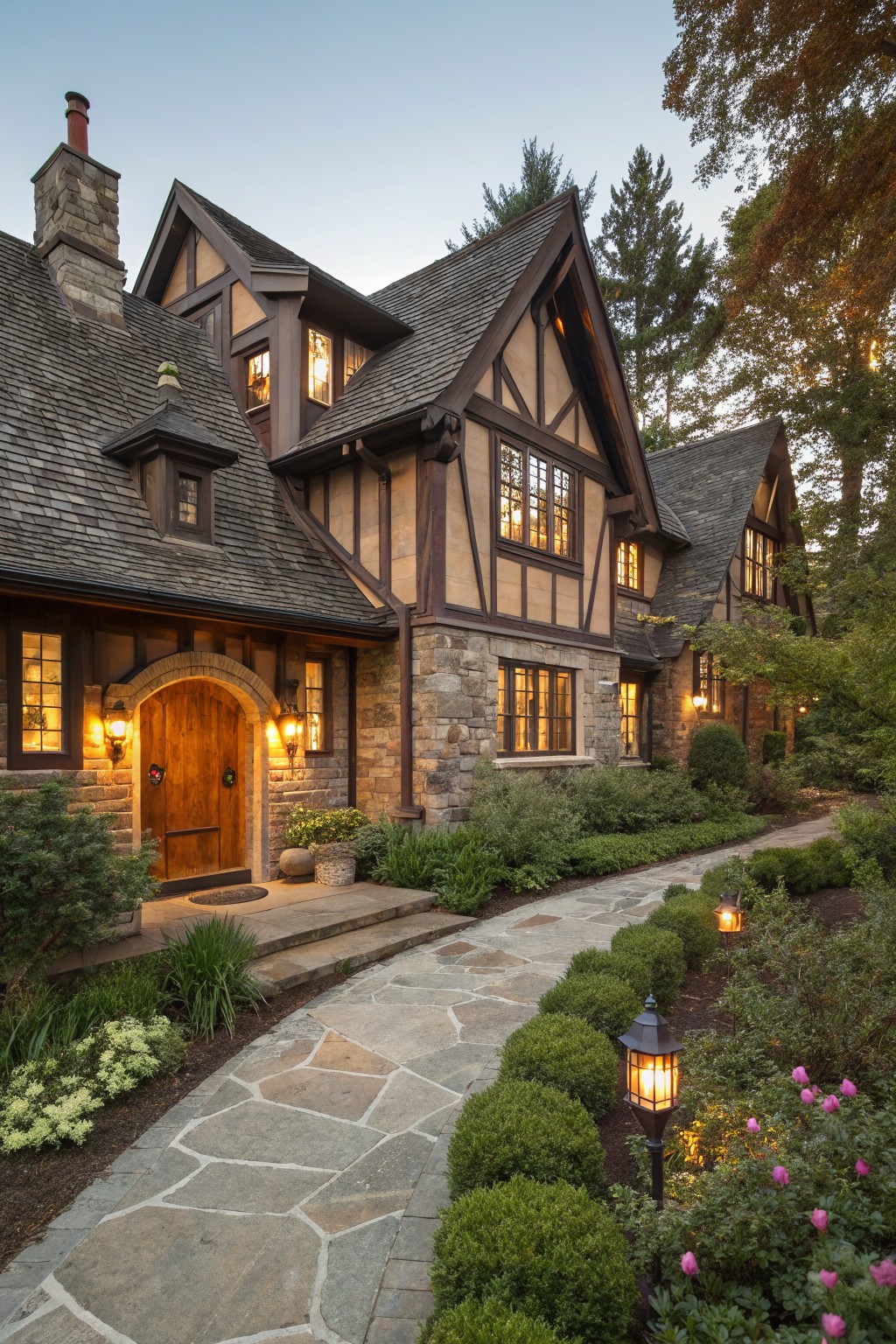 Tudor-style house exterior with dark brown half-timbering over beige stone walls, multi-gabled roof, arched wooden front door with lanterns, flagstone pathway lined with shrubs and garden lights, surrounded by trees at dusk.