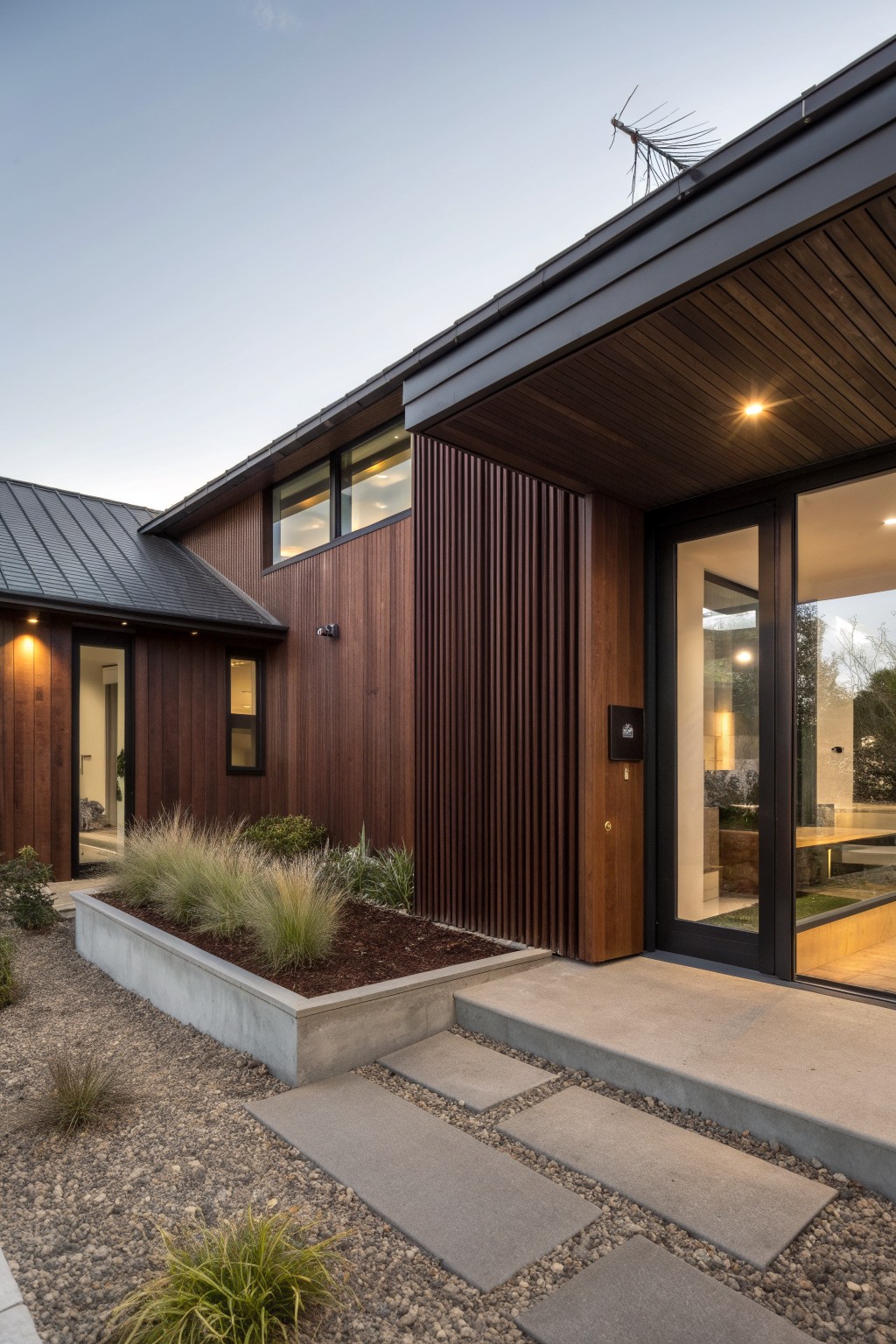 Side view of a modern house exterior with dark brown vertical wood slats beside black-framed glass entry doors, charred wood siding, standing seam metal roof, raised concrete planters filled with tall grasses, and concrete stepping stones on gravel ground.