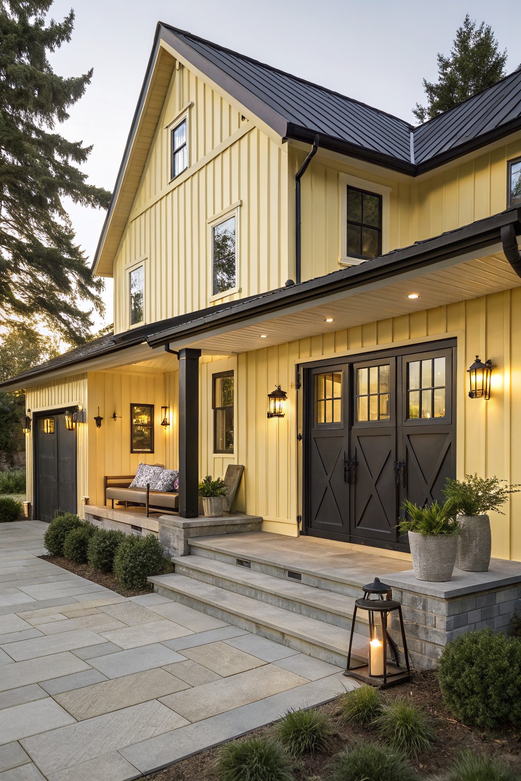 Two-story yellow board-and-batten house exterior with black metal roof, black double front doors, black garage doors, covered porch with bench, stone steps, lanterns, potted plants, and shrubs.