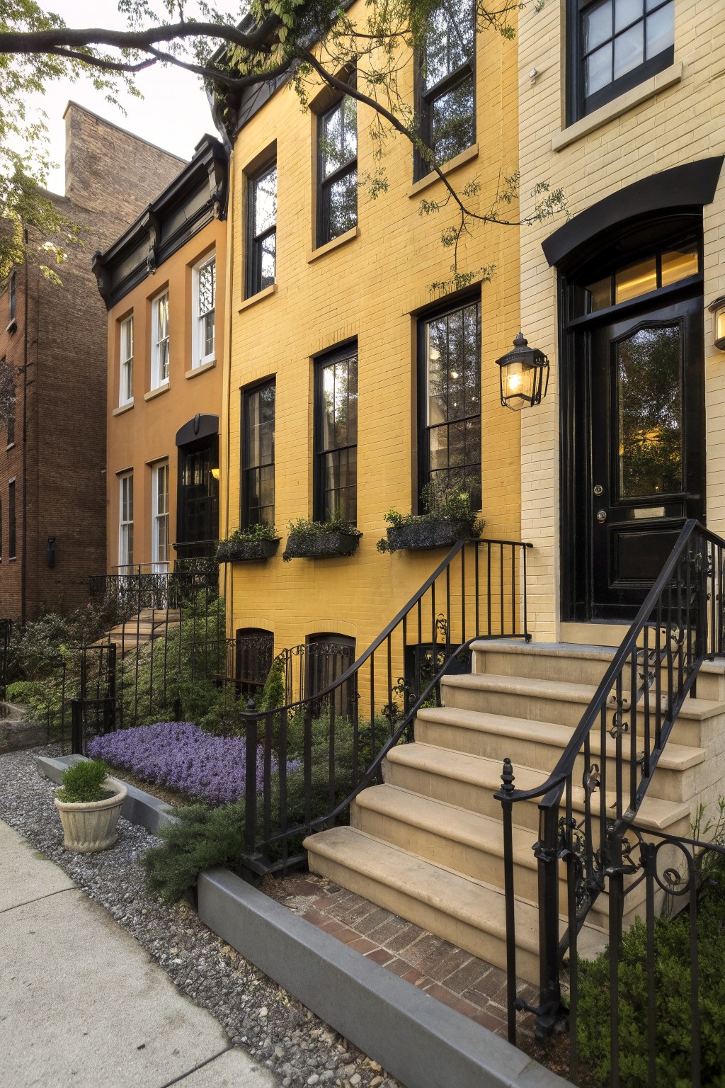 Row of three-story brick townhouses painted yellow, terracotta, and beige with black window frames, trim, and railings, plus stone entry steps and front plantings.