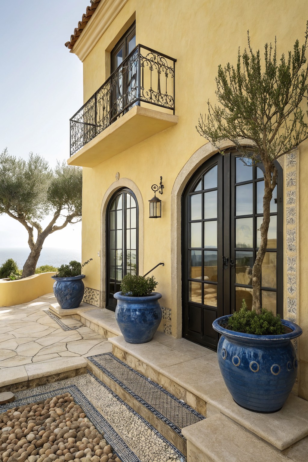 Yellow stucco house exterior with black metal arched double doors, wrought iron balcony above, blue ceramic planters with olive trees and shrubs, stone steps, and pebble mosaic path.