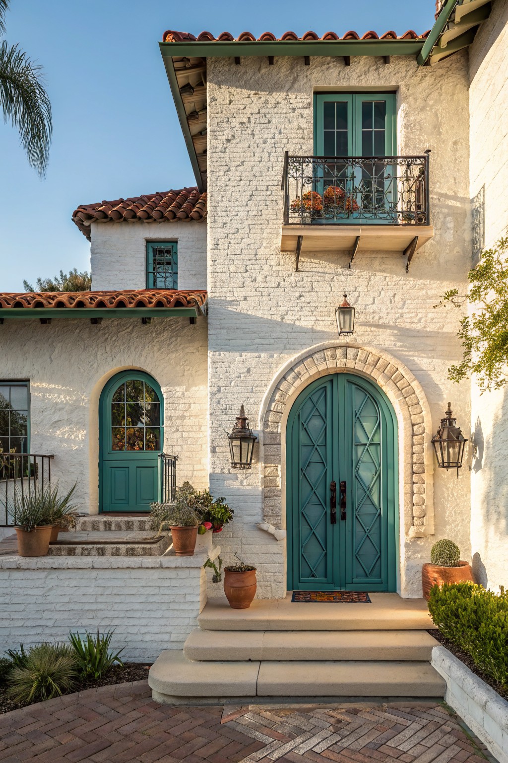 White stucco house exterior with arched teal double front doors flanked by lanterns, matching teal windows and side door, terracotta tile roof, potted plants on steps, and brick pathway.