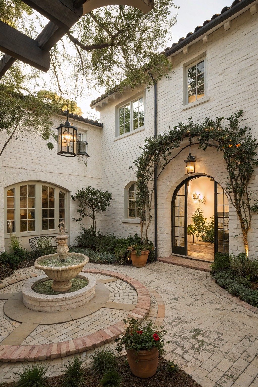 White brick house exterior with enclosed courtyard centered on a stone fountain, arched glass entryway framed by climbing vines and lanterns, potted plants, trees overhead, and stone paving path.