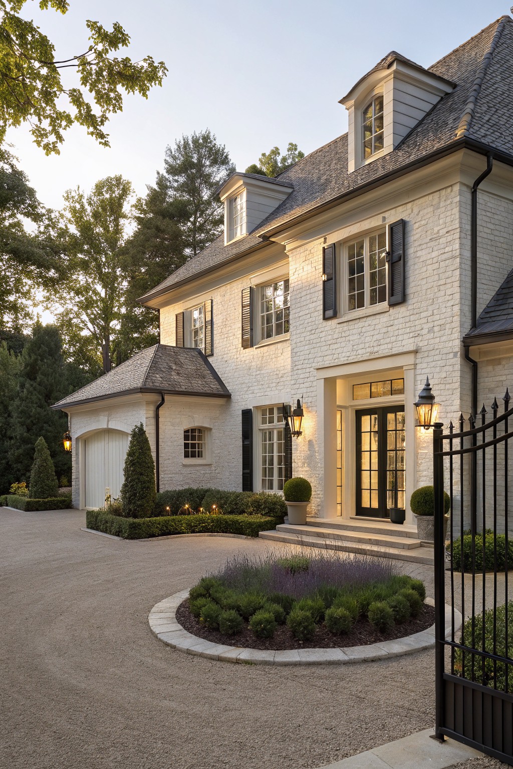 Two-story white brick house exterior featuring black shutters, multi-pane windows, attached garage with wood door, curved gravel driveway with central circular planting bed, black iron gate, entry steps, and flanked lanterns.