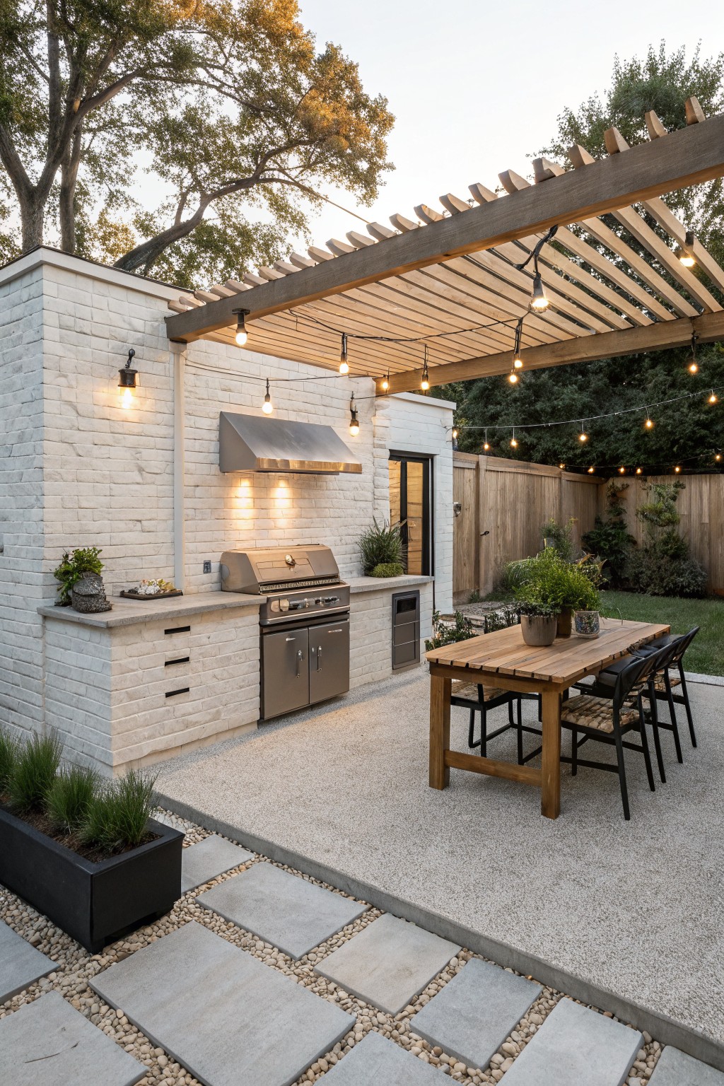Outdoor kitchen with white brick walls and counters, stainless steel grill and hood, under wooden pergola with string lights, next to wooden dining table and chairs on gravel patio surrounded by plants and grass.