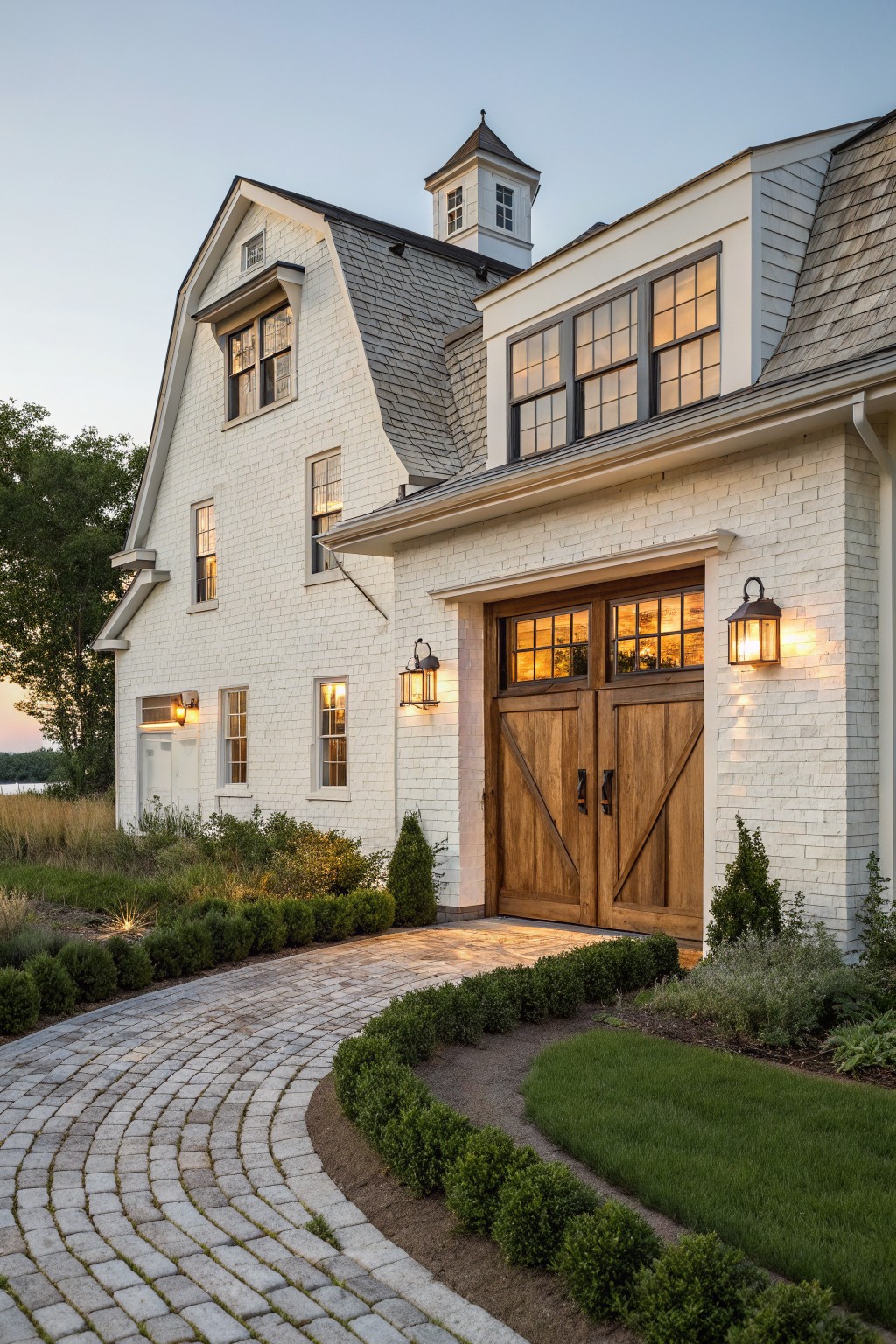 White brick house exterior featuring a gambrel roof, cupola, large wooden double garage doors with glass panels, brick pathway, boxwood hedges, and landscape lighting at dusk.