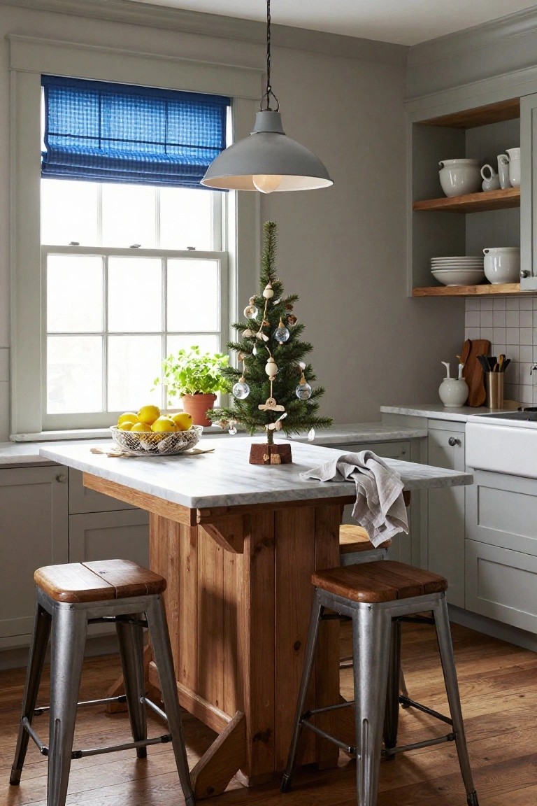 Coastal kitchen corner with gray cabinets, wooden island on metal stools, small decorated Christmas tree and lemons on marble countertop, blue roman shade at window, and pendant light overhead.