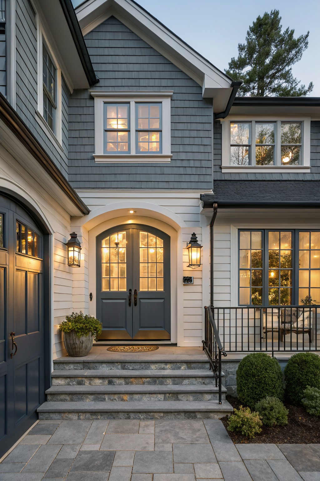 Gray shingled house exterior with white-trimmed arched entry featuring navy double doors, lanterns on each side, navy arched garage doors, stone steps, bluestone pavers, and boxed shrubs.