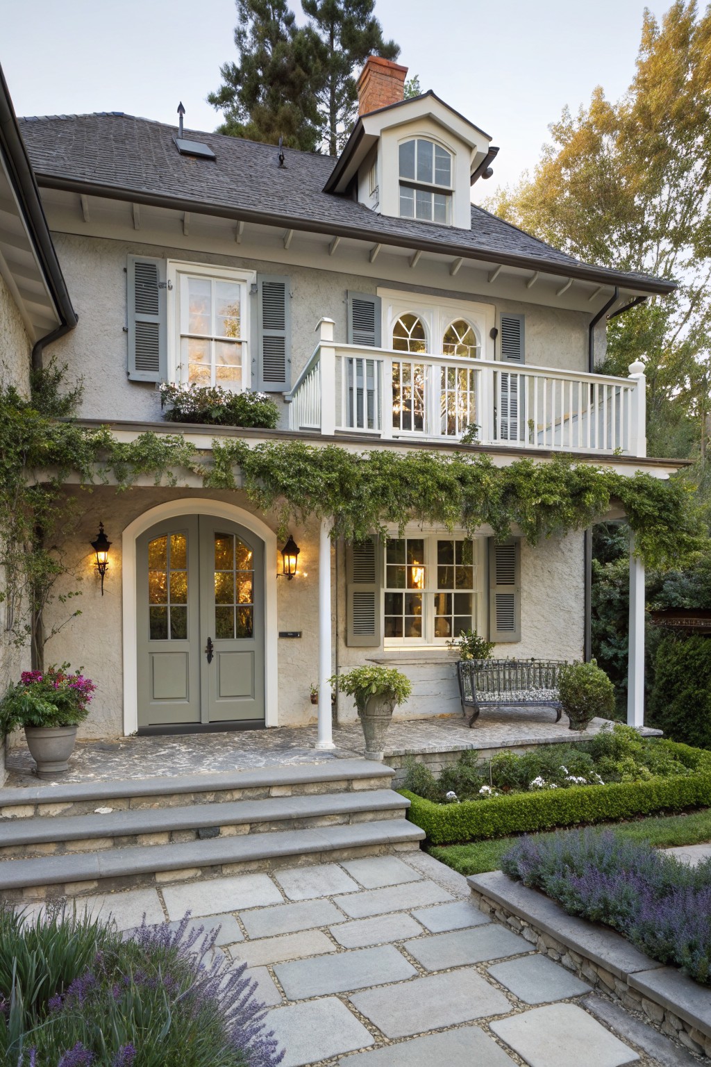 Two-story gray stucco house exterior featuring an arched double door entryway covered in climbing vines, white-trimmed windows, upper balcony, stone steps, potted plants, and lavender garden borders.