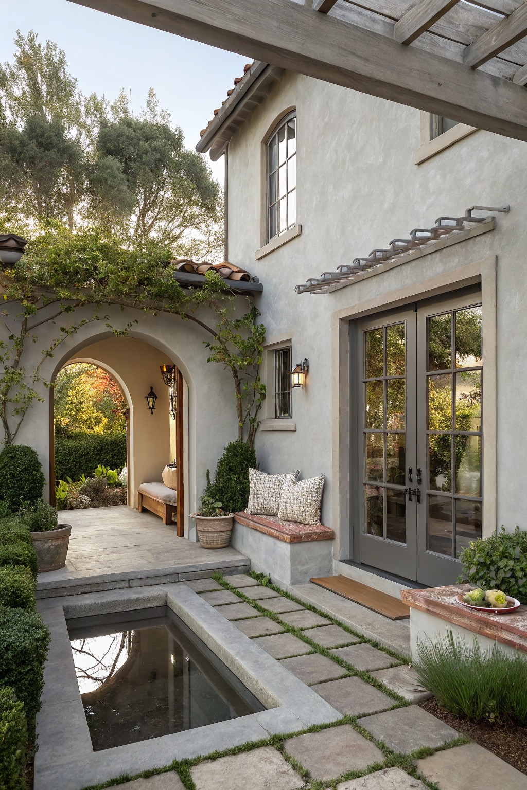 Gray stucco courtyard with a small square reflecting pond in concrete pavers, built-in bench with cushions on a low wall, French doors on house exterior, arched vine-covered entryway, potted plants, and grasses.