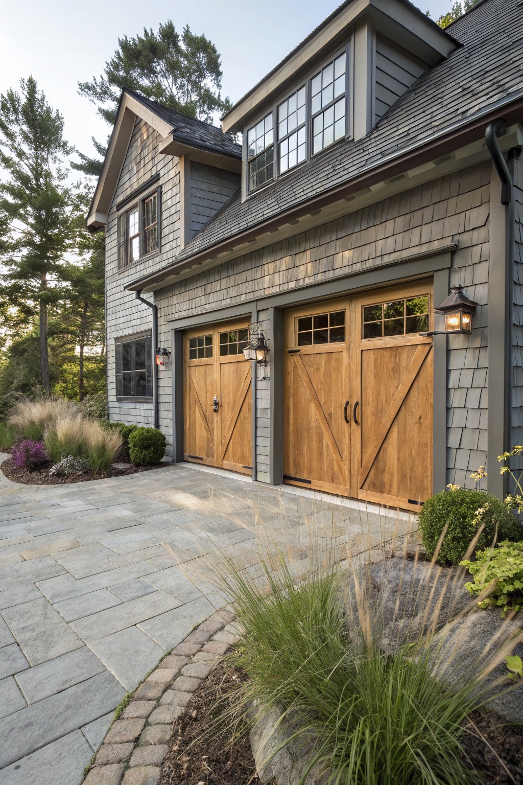 Gray shingle house exterior featuring double wooden garage doors with glass panels and lanterns, next to a stone paver driveway edged with grasses and shrubs.