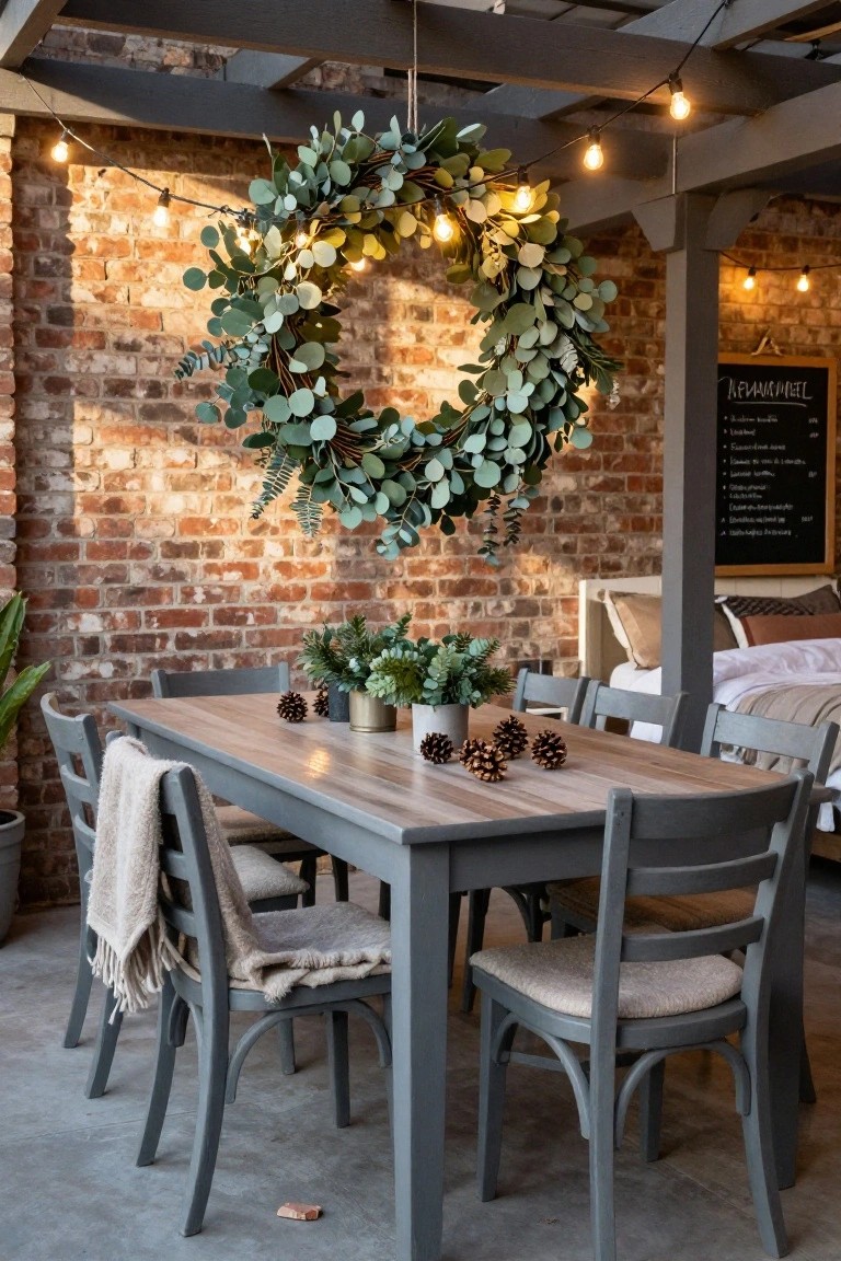 Outdoor pergola with string lights and large illuminated eucalyptus wreath hanging above a gray wooden dining table and chairs, against a brick wall with plants and a chalkboard.