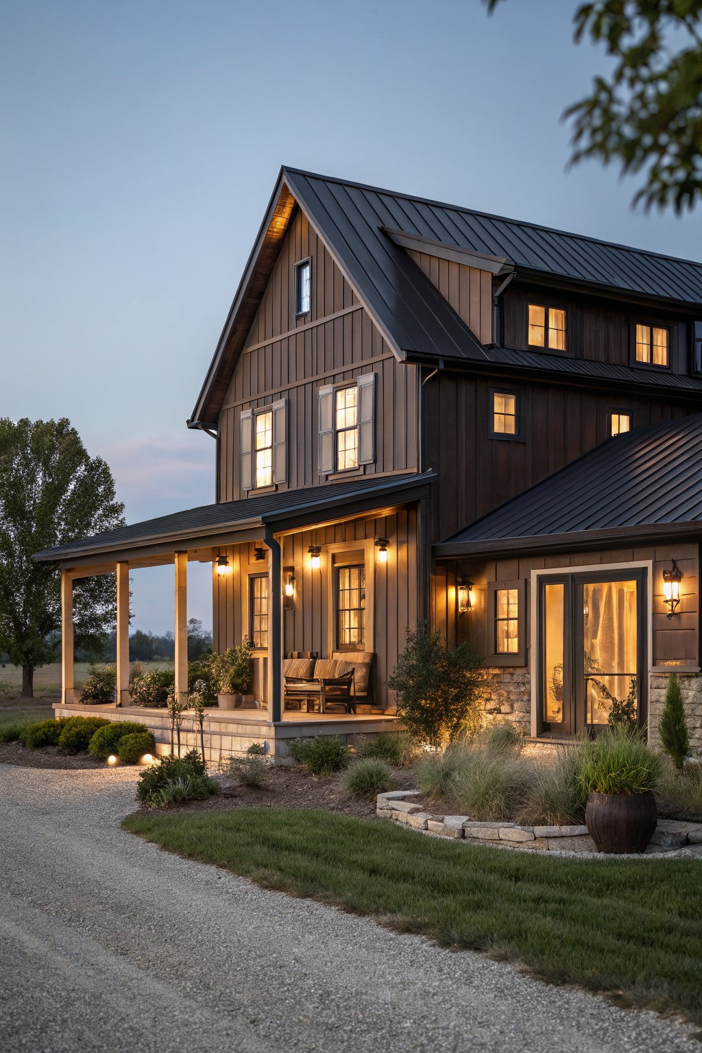 A two-story house with dark brown board-and-batten siding, black standing-seam metal roof, wraparound porch with lanterns, stone accents, landscaping, and gravel driveway at dusk.