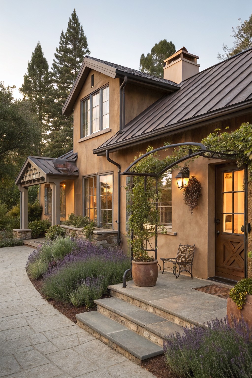 Brown stucco house exterior with dark metal roof, vine-covered arched entryway, wooden door, lavender plants along stone path and steps, and tall trees in background.