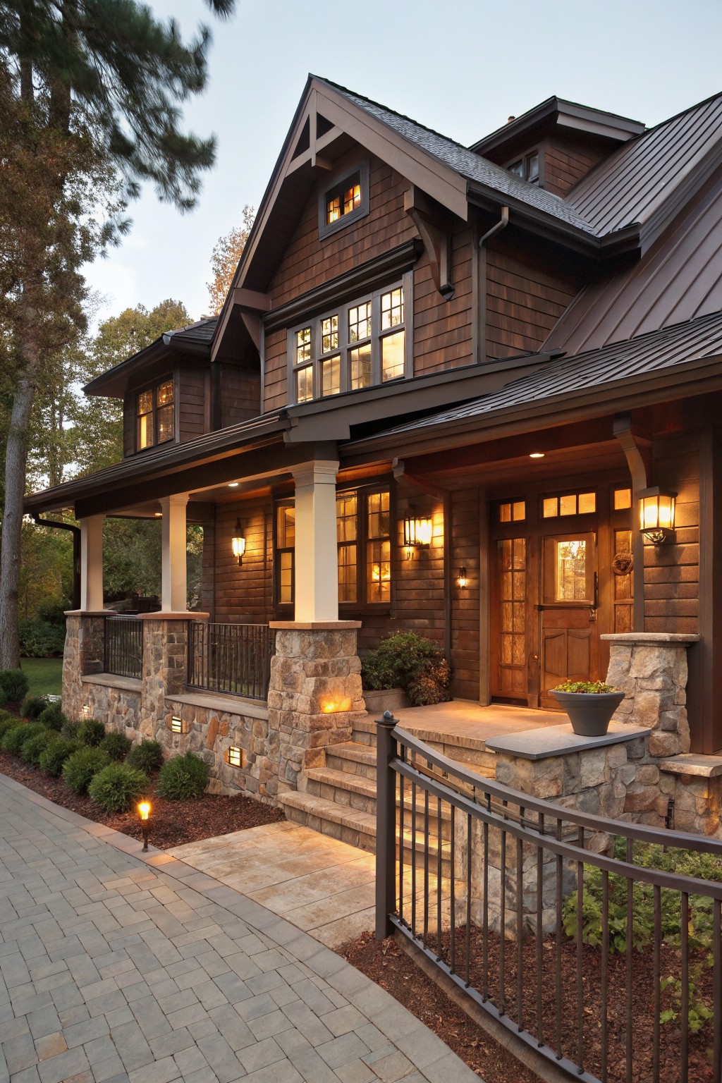 A two-story house with brown shingle siding, dark standing seam metal roof, wraparound porch supported by stone pillars, landscape lighting, and surrounding trees and shrubs at dusk.