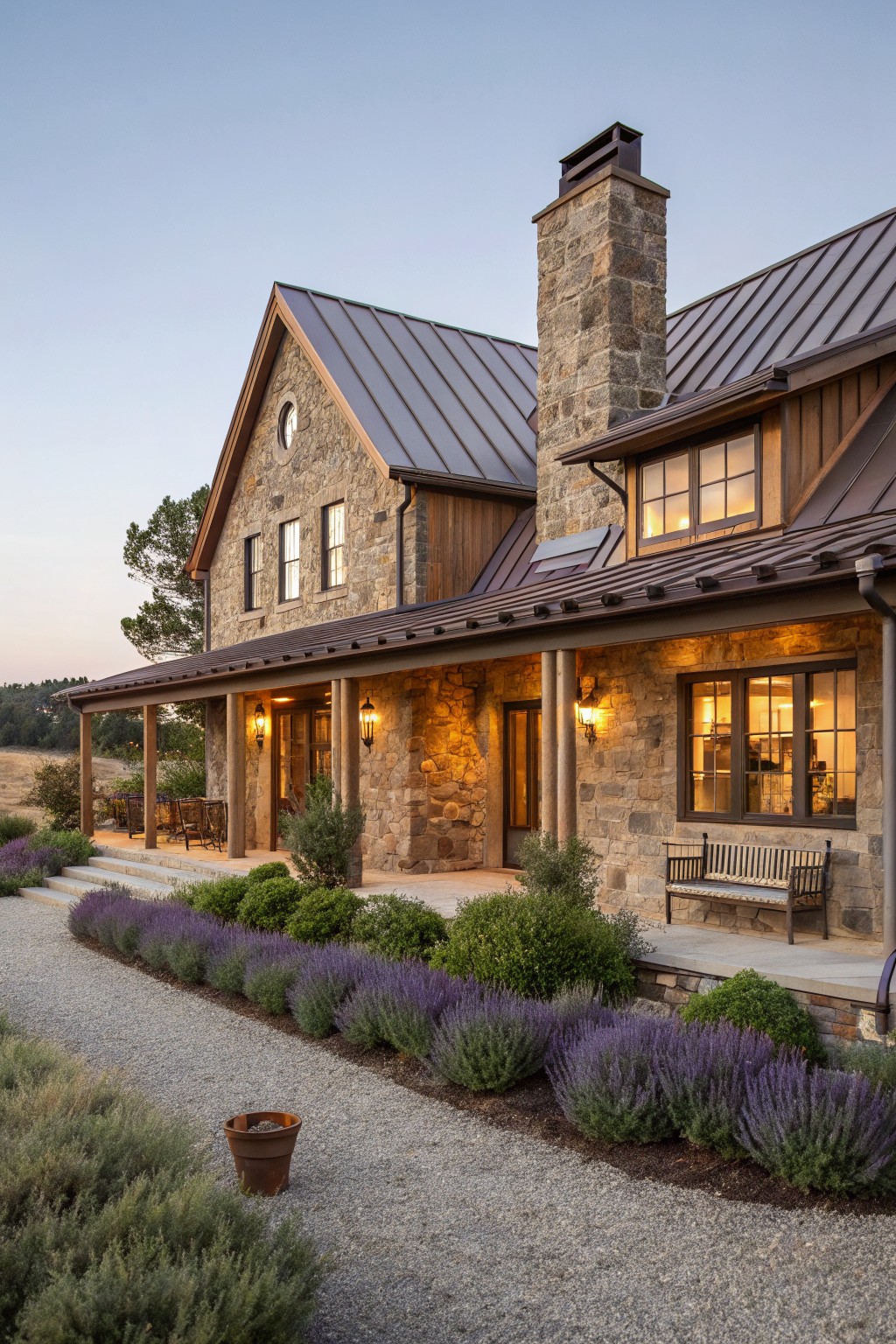 Two-story house exterior featuring stacked stone walls, wood porch with columns, brown metal standing seam roof, large windows, and gravel path lined with lavender plants at dusk.