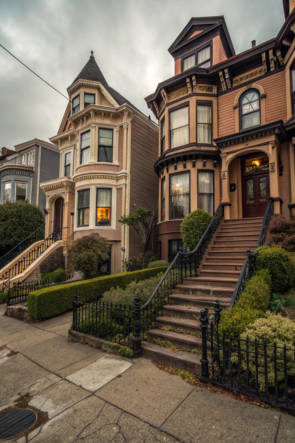 Two brown Victorian houses side by side on a city street, with ornate trim, bay windows, steep stairs flanked by black wrought iron railings, hedges, and overcast skies.
