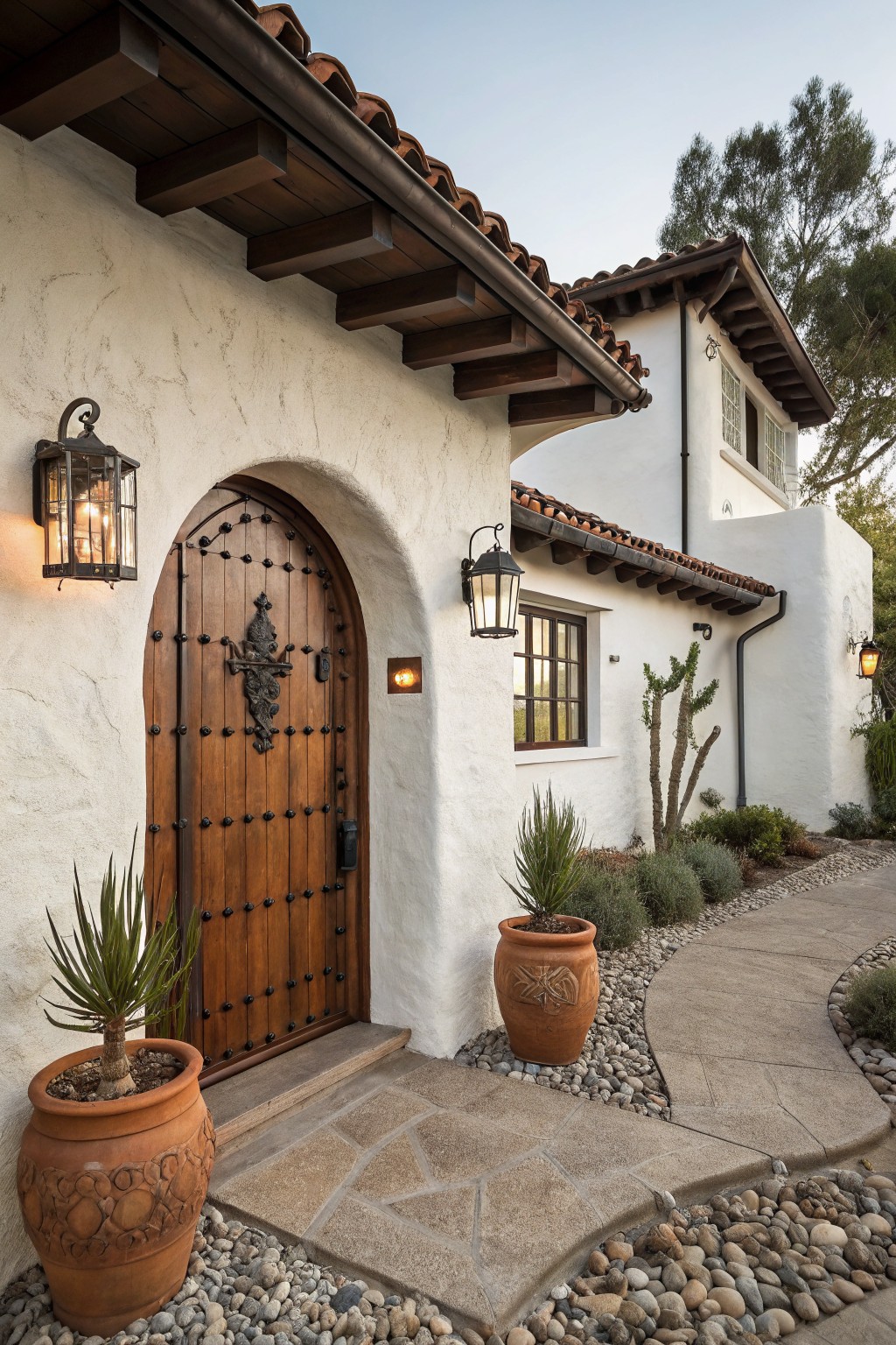 White stucco house exterior with arched dark wooden front door featuring iron studs and handle, flanked by lanterns and large terracotta pots with agave plants, terracotta tile roof, and curved stone pathway.
