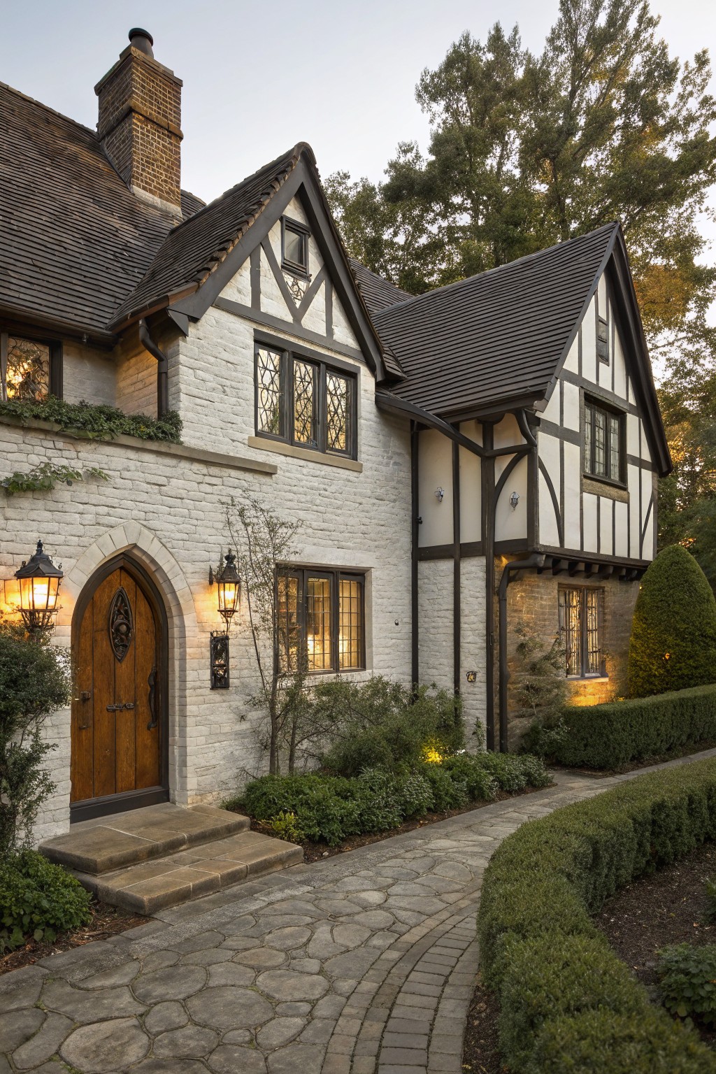 Tudor Revival house exterior featuring white brick walls, dark timber framing, dark shingled gabled roofs, arched wooden front door with lanterns, stone steps, curved flagstone pathway, and low boxwood hedges.