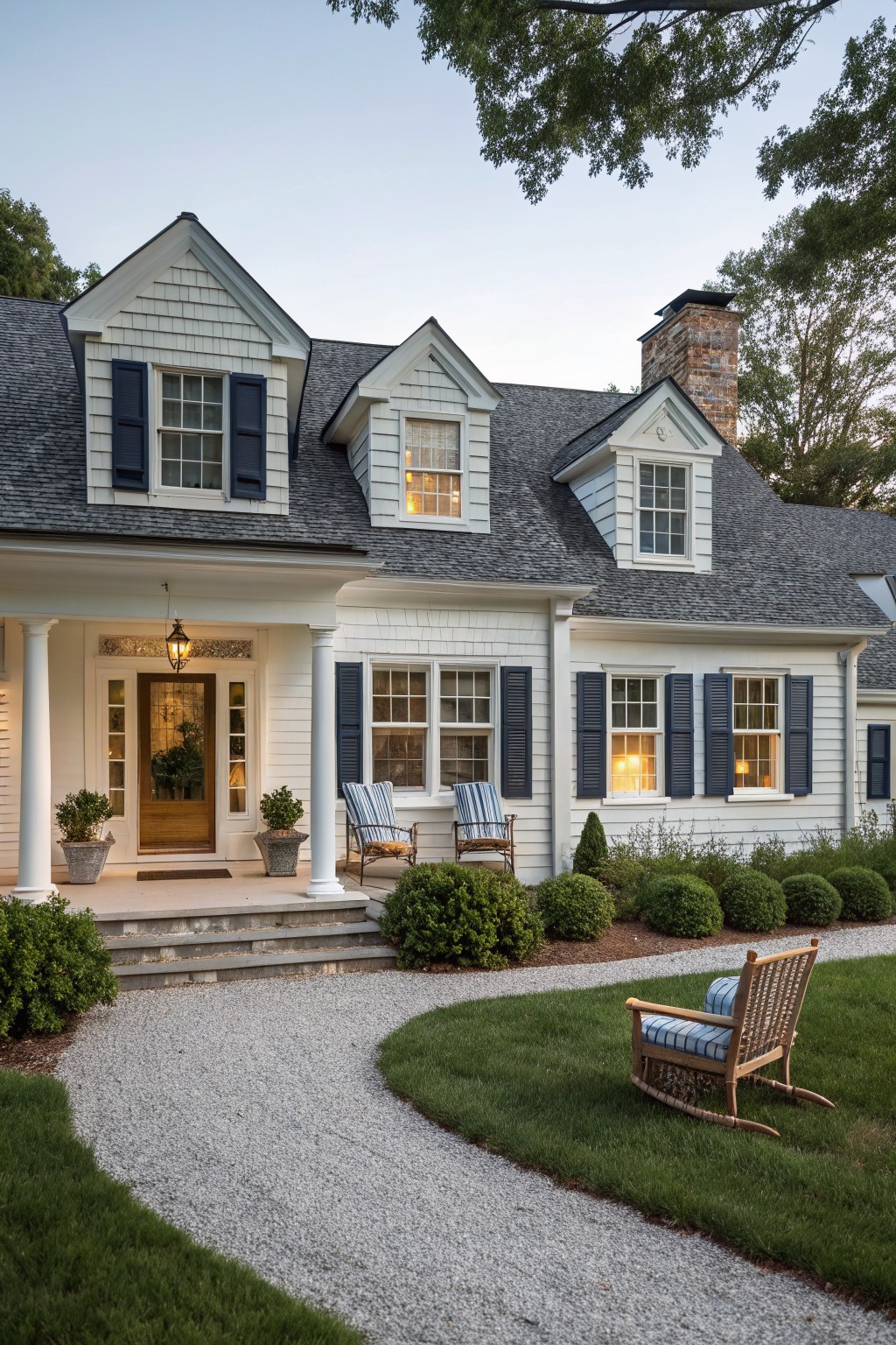 White clapboard house exterior with navy shutters, columned front porch, potted plants, Adirondack chairs, gravel pathway, and surrounding shrubs at dusk.