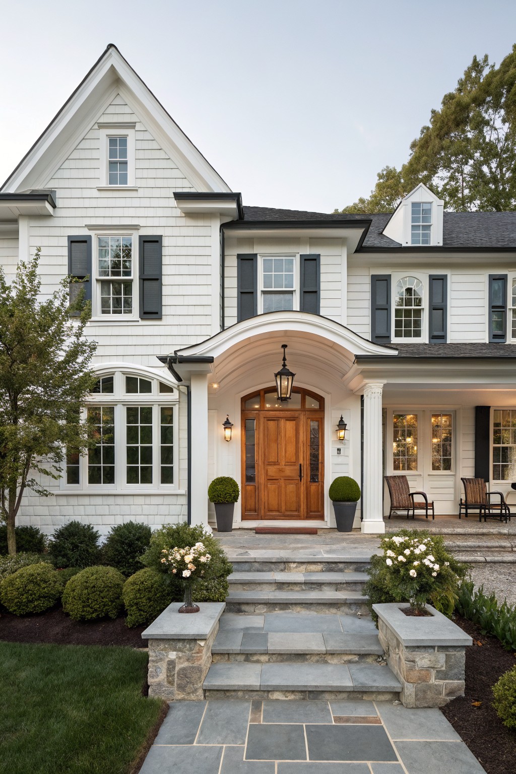 Front exterior of a two-story white shingle house with black shutters, arched porch entry featuring a wooden door and lanterns, stone steps flanked by shrubs, and a landscaped yard.