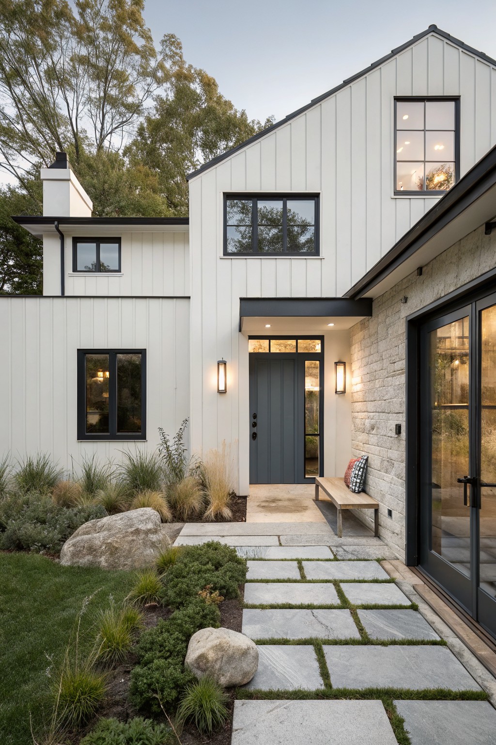 White board-and-batten sided house exterior with black-framed windows, dark gray front door flanked by lanterns, stone entry wall, paver pathway, boulders, and grasses leading to the front door.