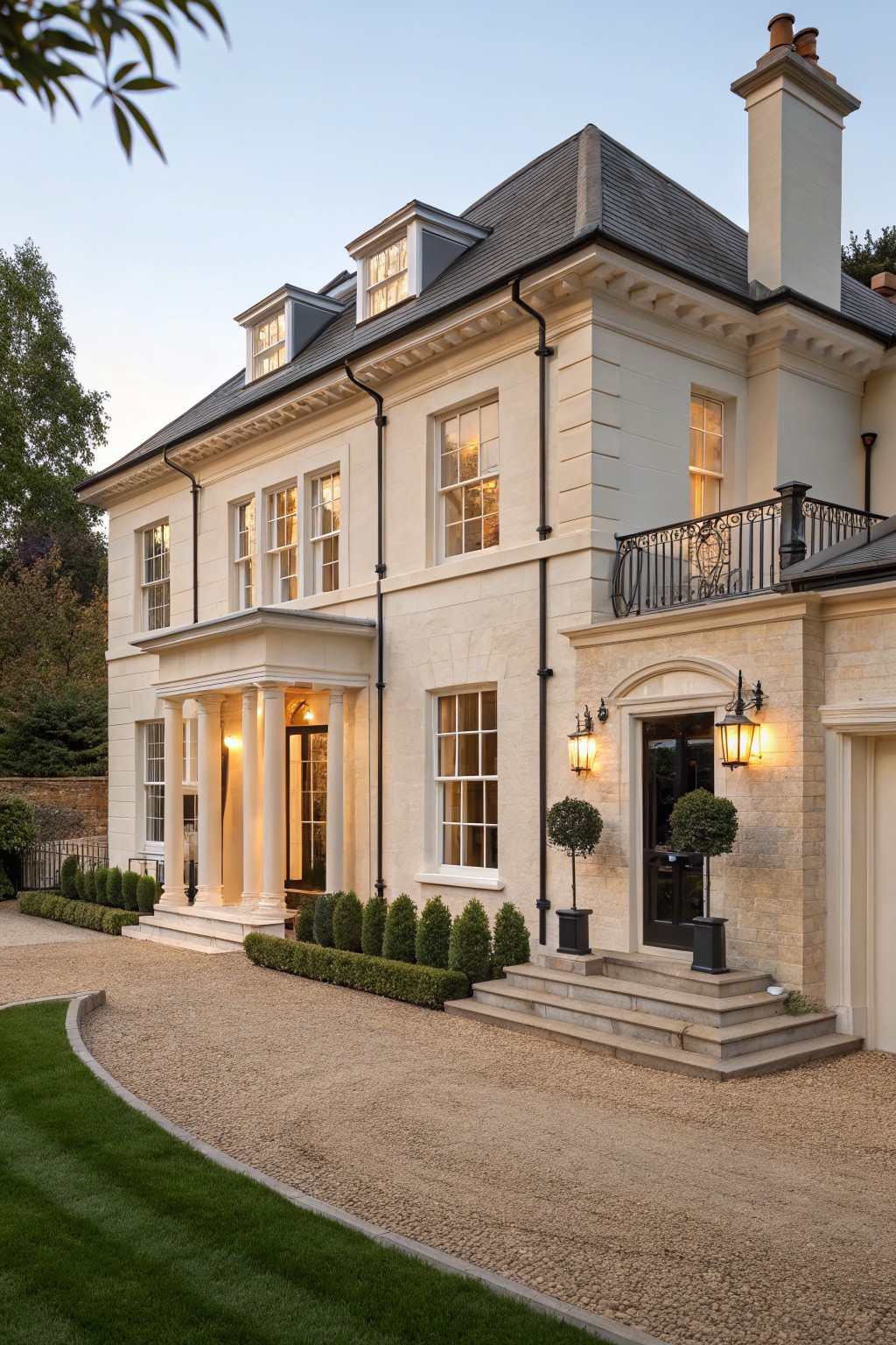 Two-story light beige stone house with slate roof, classical columned portico entry, black doors and lanterns, boxwood shrubs, and gravel driveway edged by grass.