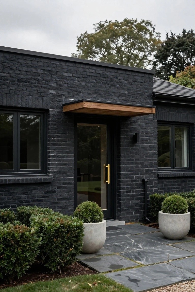 Front view of a modern house featuring black brick walls, a wooden canopy over the glass entry door with gold handle, black-framed windows, potted boxwood shrubs, and a slate stone pathway.