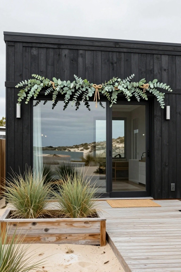 Black wooden house exterior featuring a long eucalyptus garland draped over large sliding glass doors, with grass planters in wooden boxes on a deck near sand dunes.