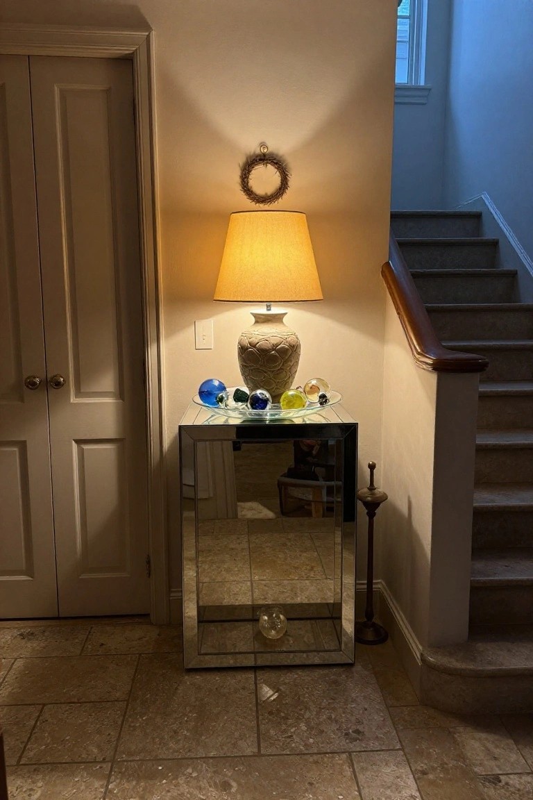 Mirrored console table in an entryway holding colorful glass Christmas ornaments on a plate next to a ceramic vase, with a lamp nearby, double doors, and carpeted stairs.