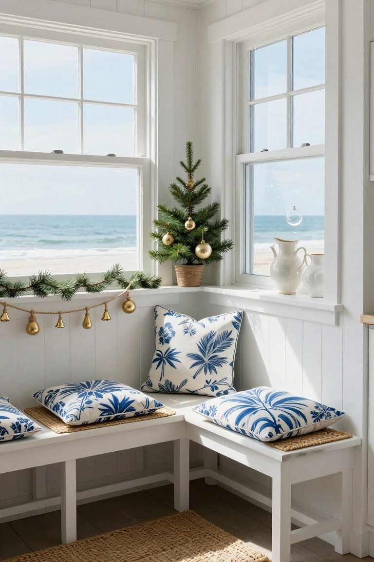 White paneled corner room with window seat, blue palm-print pillows, small potted Christmas tree, gold garland with bells and ornaments on windowsill overlooking beach and ocean.