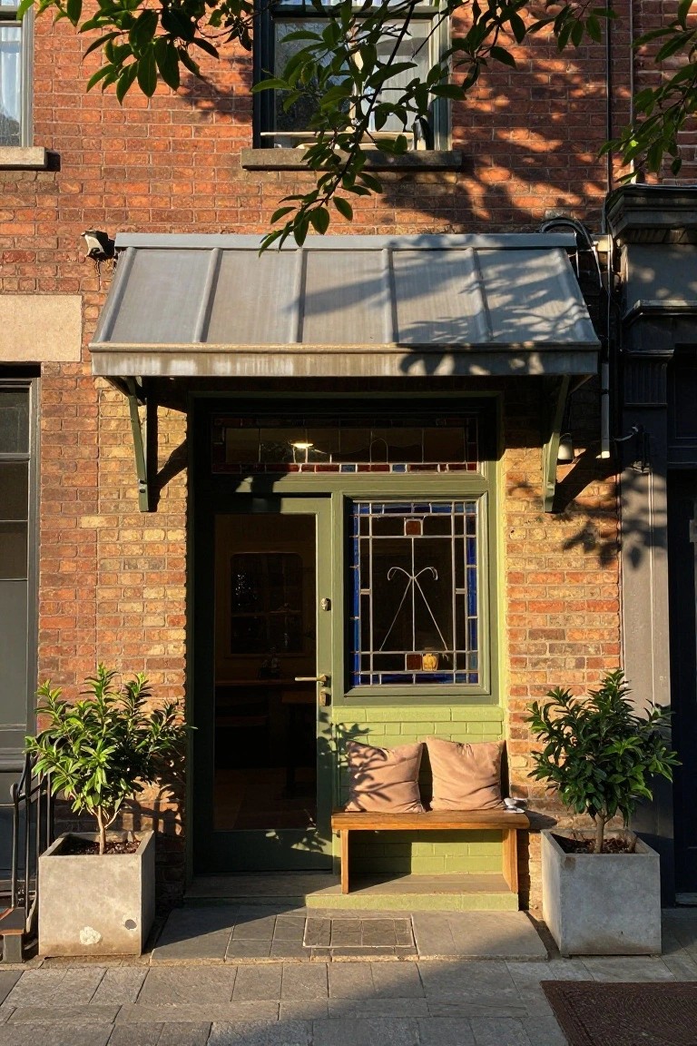 Brick house front entrance with green door featuring blue and multicolored stained glass window, wooden bench with beige cushions, potted green plants in concrete pots on either side, metal awning overhead, and tree shadows in late afternoon light.