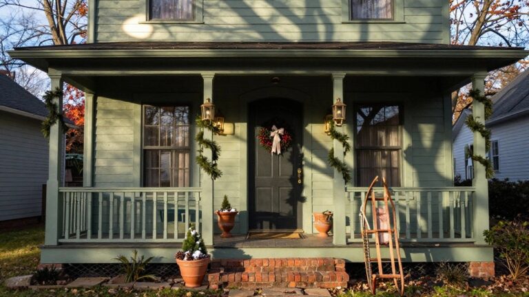 Sage green house with white porch columns wrapped in evergreen garlands, dark green front door with berry wreath, flanked by lanterns, potted plants with snow, pinecone baskets, and wooden rocking sled on brick steps.