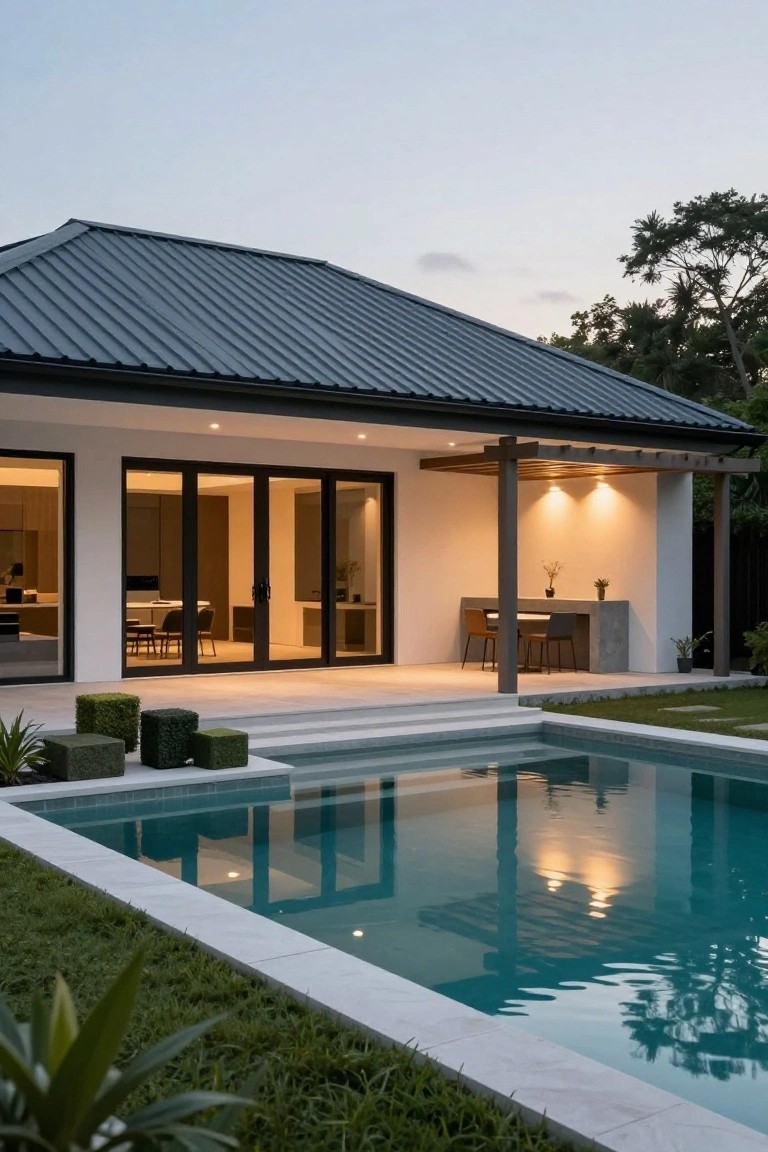 Exterior view of a modern white stucco house with dark gray ribbed metal roof, large black-framed glass doors opening onto a patio with concrete bar counter and stools under a wooden pergola, steps descending to turquoise pool edged in white stone, surrounded by green lawn and tropical plants at dusk.
