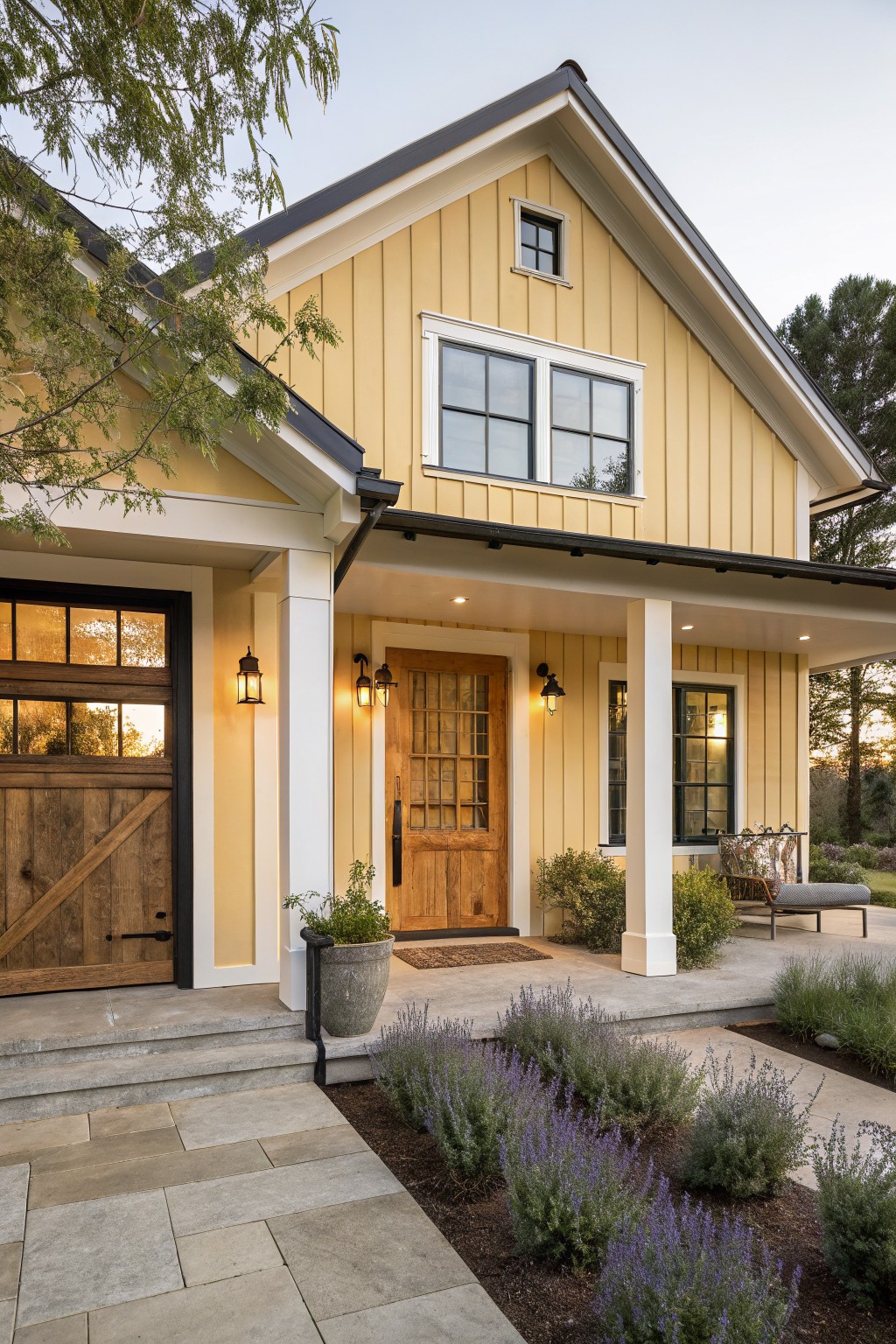 A two-story yellow board-and-batten house with white trim, black roof and gutters, covered front porch with wooden barn-style garage door and paneled entry door, flanked by white columns and lanterns, stone steps leading to a pathway edged with lavender plants.