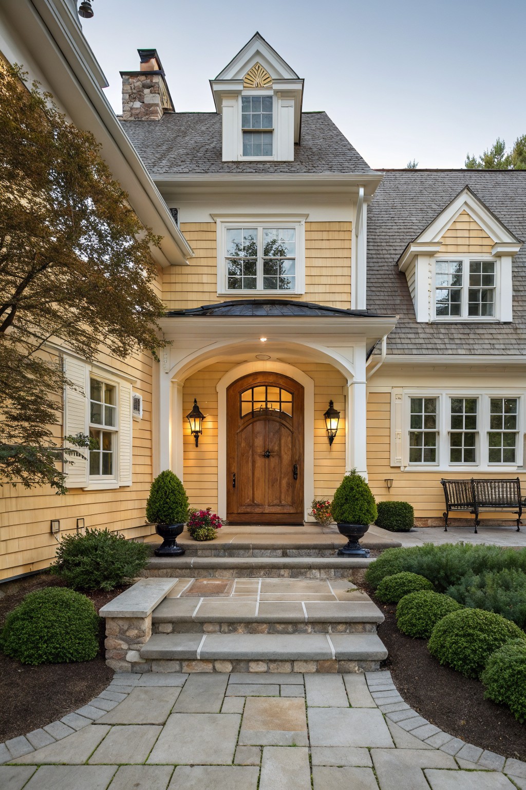 Front view of a two-story yellow shingle house with white trim, an arched wooden entry door flanked by lanterns, stone steps, potted plants, and low shrubs.
