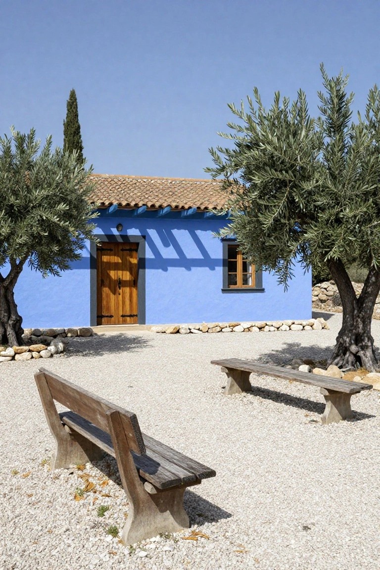 Blue stucco house with terracotta tile roof, dark wooden double door entry, small side window, flanked by two olive trees, gravel courtyard with two wooden benches and stone borders.