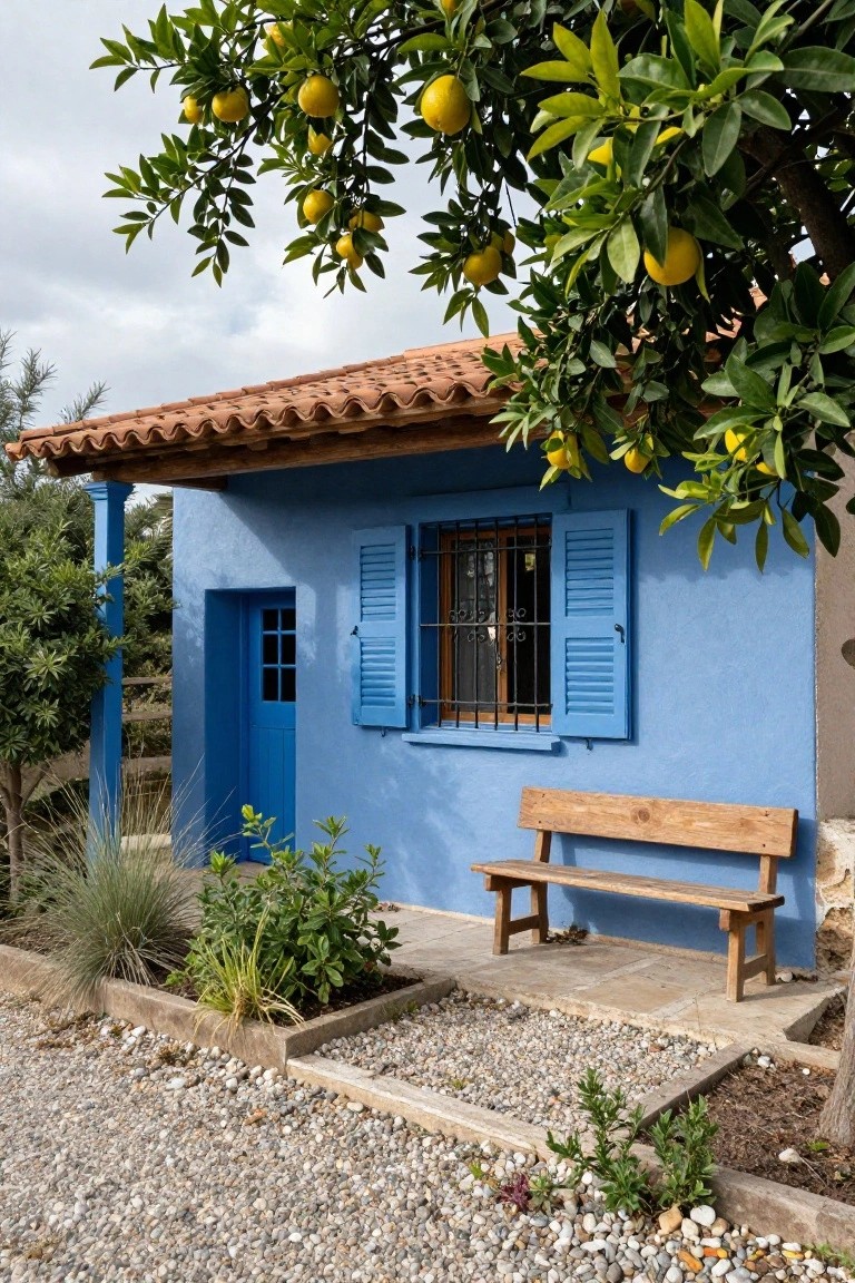 Small blue stucco house with terracotta tile roof, blue door, barred window with blue shutters, wooden bench on gravel path beside it, overhanging lemon tree, and surrounding plants and shrubs.