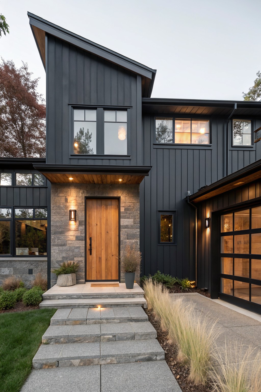 Modern house exterior with dark gray vertical board-and-batten siding, asymmetrical sloped roof, wooden front door in stone surround, large glass garage door, concrete pathway, and low landscaping along stone steps.